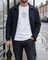 Man at a rainy bus stop wearing the British Weather Club tee in White under an open navy jacket